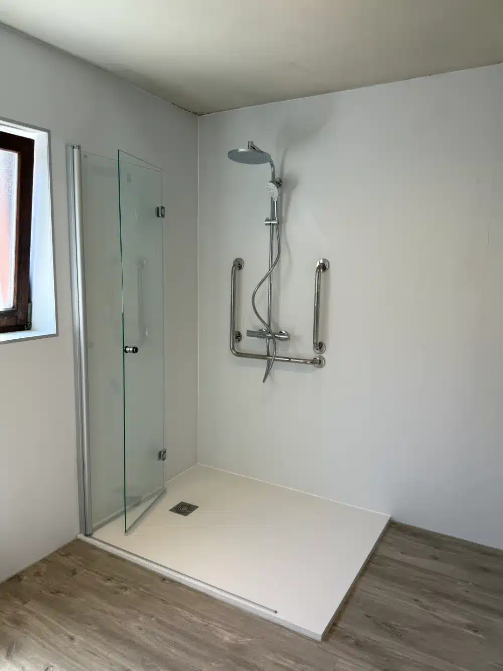 Douche senior de plain-pied avec double barre d'appui coudée, pommeau de tête et porte battante-pliante en verre, réalisation Hyseco